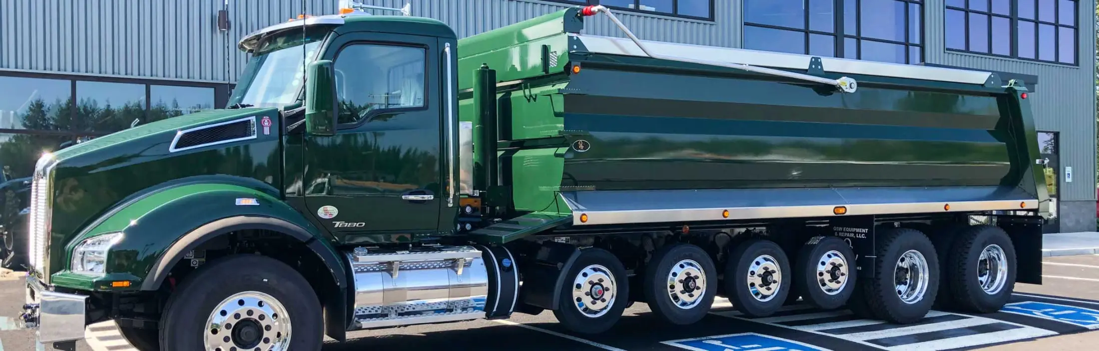 A green tri-axle dump truck parked outside a depot